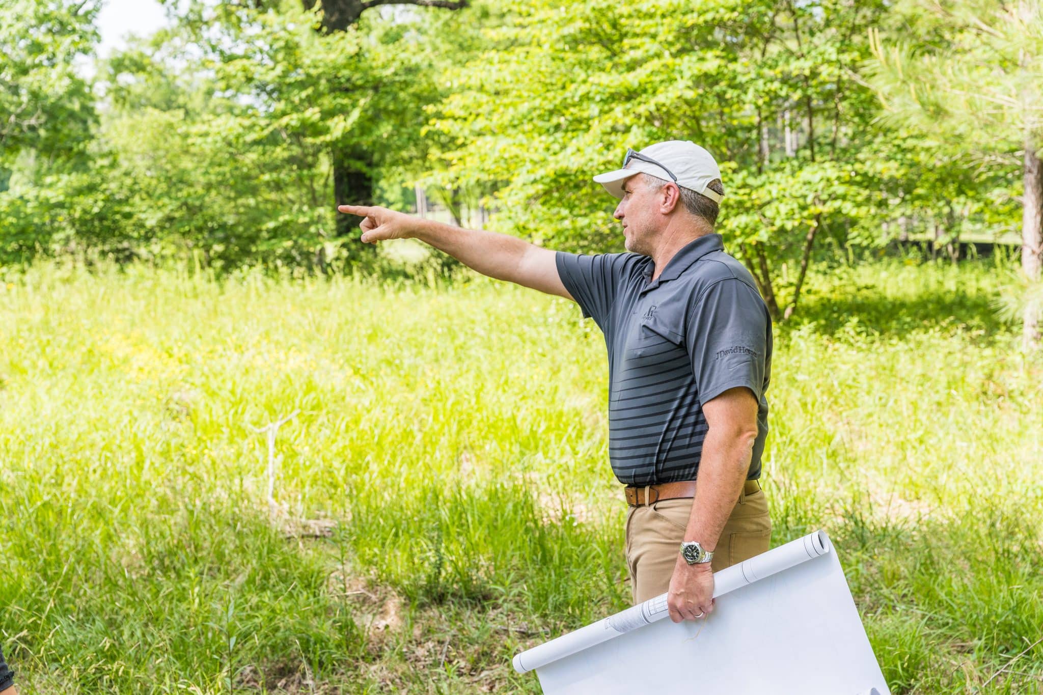 A man holding custom home plans stands in a plot of land for sale in Birmingham and points at distant feature