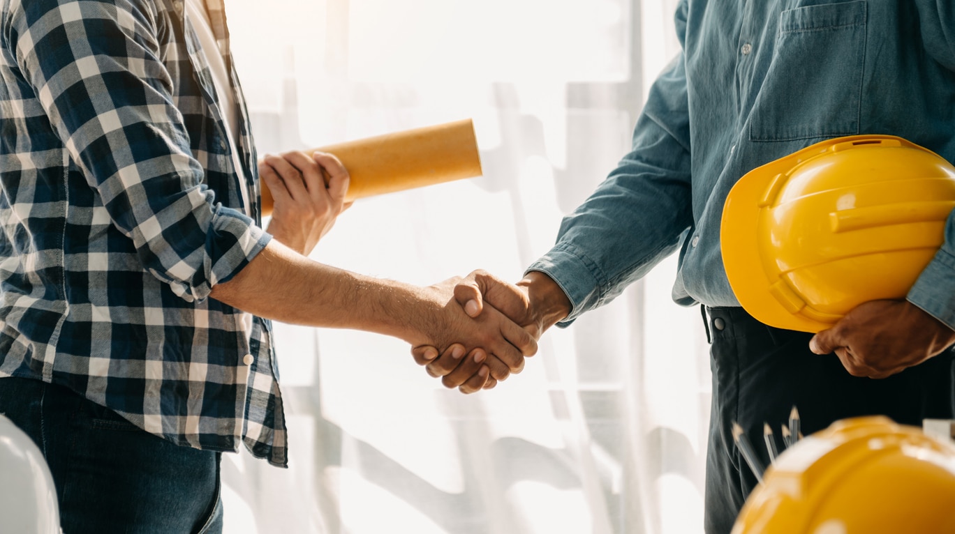 A home builder and homeowner shake hands with plans and construction hats in hand.