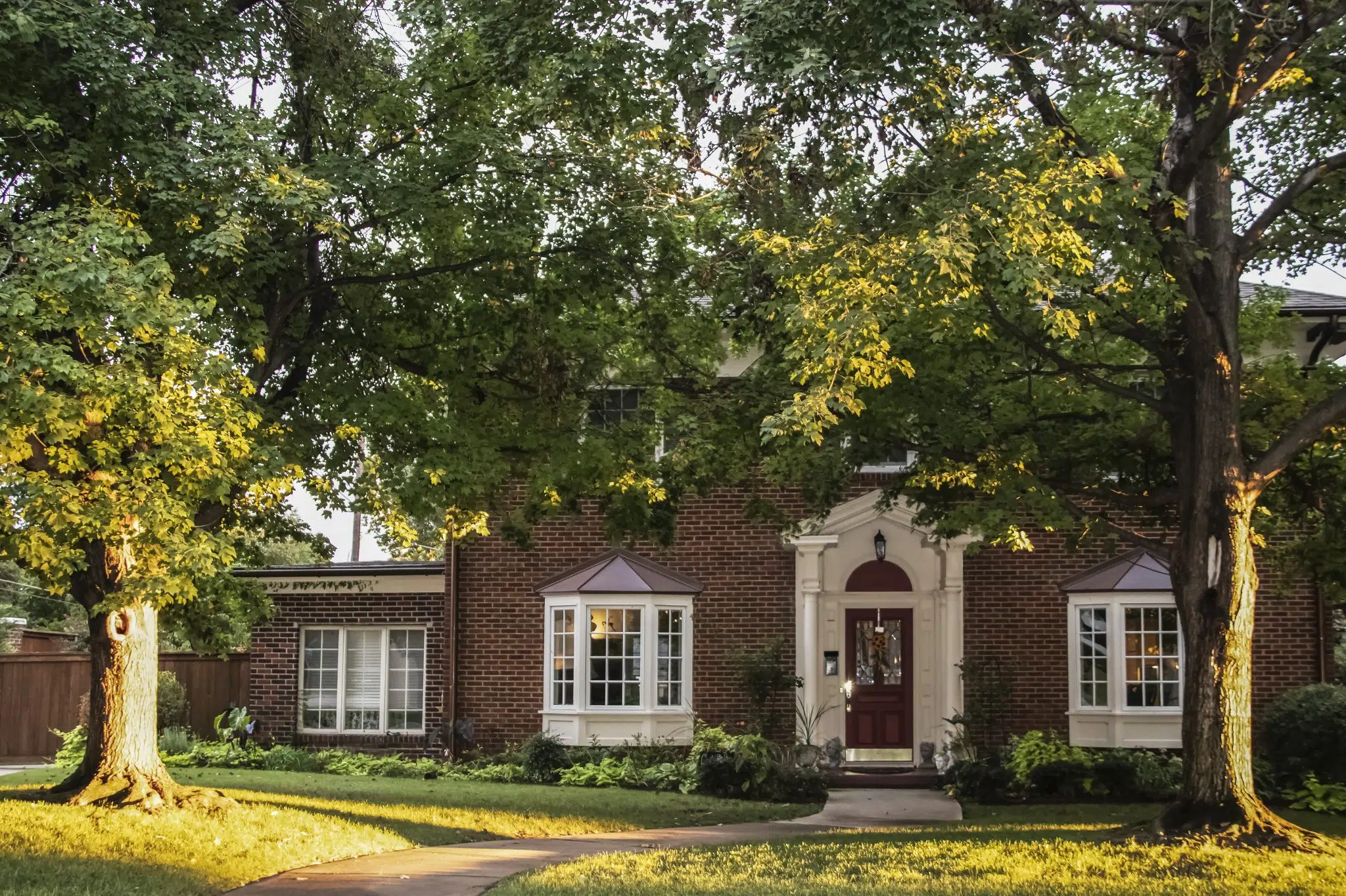 Golden Hour on the maple trees in front of traditional brick house with columns and bay windows - light stretching across front yard and up tree trunks.