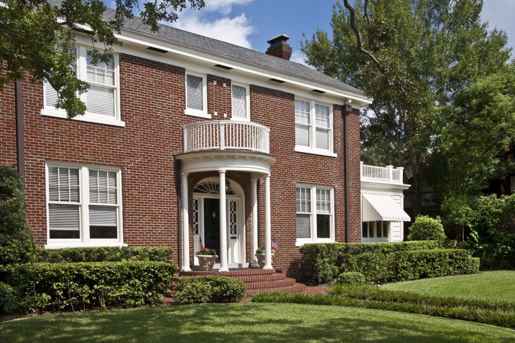 Traditional brick home with front steps, portico, front lawn, bushes, balcony and oak trees. Upscale. Two American flags on porch. Federal style architecture.