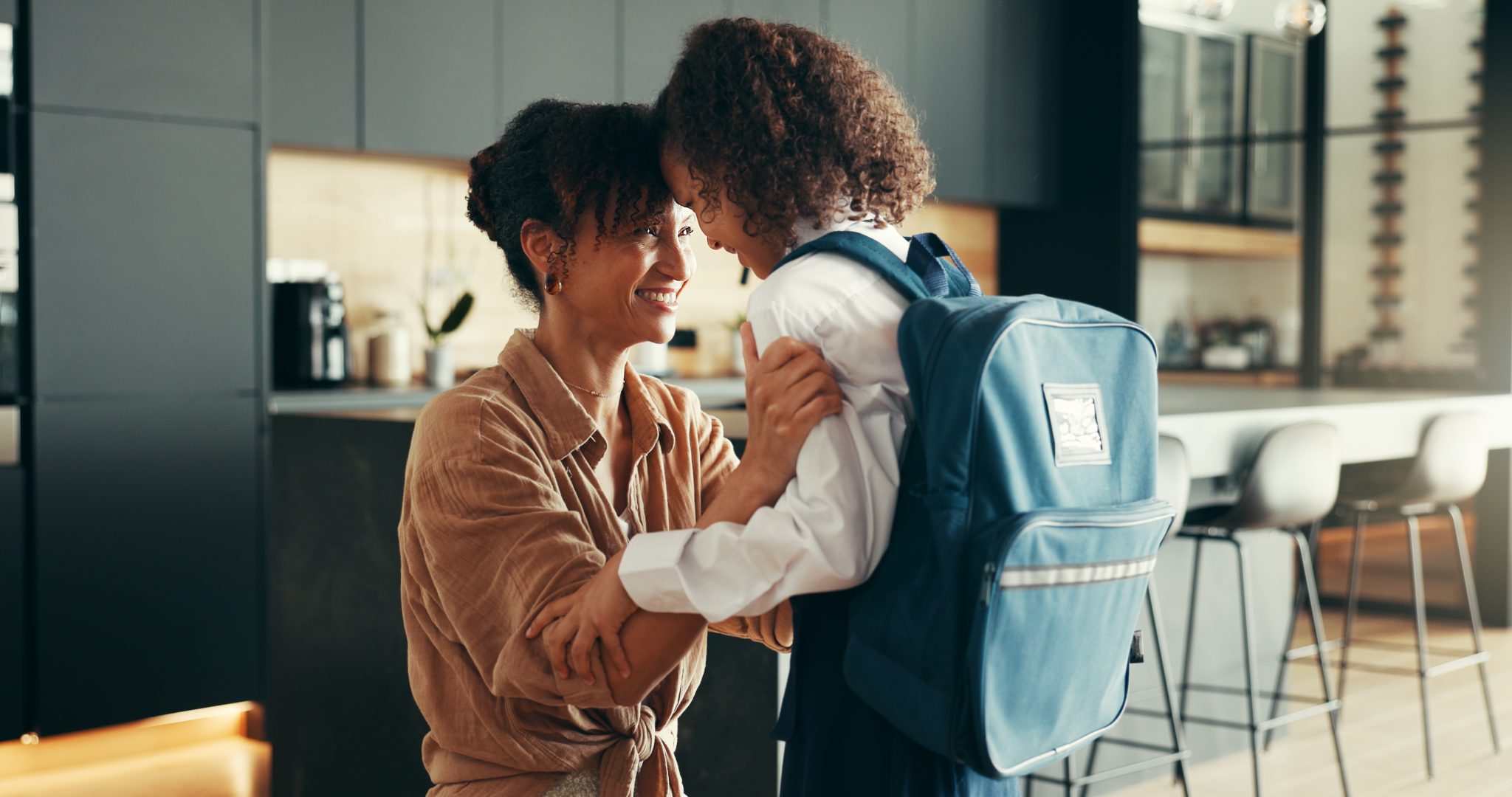 A mom and a young girl with a backpack at home in their kitchen, touching foreheads and smiling as they get ready with their back-to-school routine.