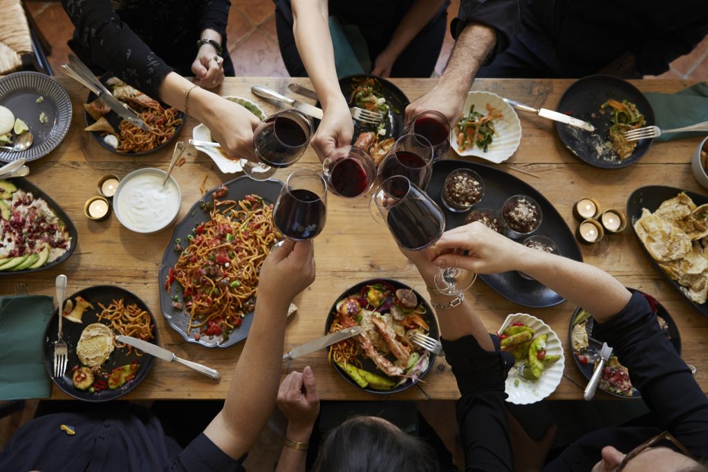 Aerial view of friends gathered around a table, enjoying different dishes and toasting glasses of wine.
