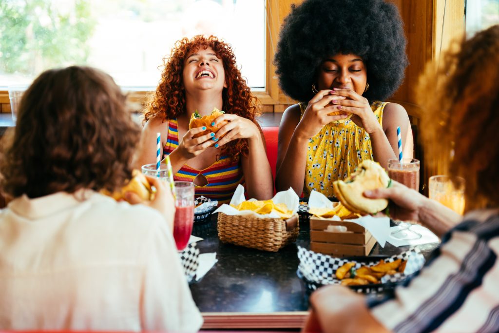 A group of women at a restaurant laughing and enjoying burgers, fries, and milkshakes.