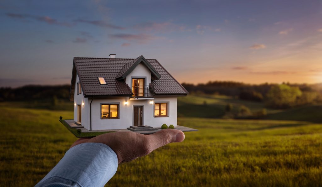 Male hand holding a model of a new dream house in the foreground of an empty field, representing custom home building and land investment. 