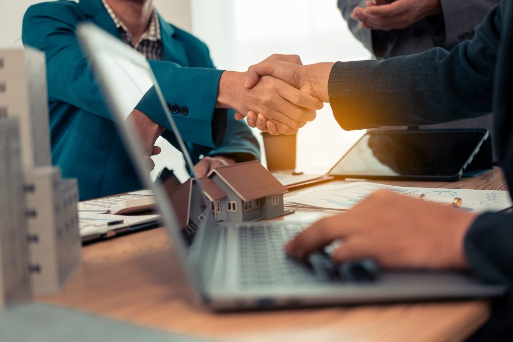 Close-up of a handshake between a real estate agent and a land buyer with a small house plan in the background.