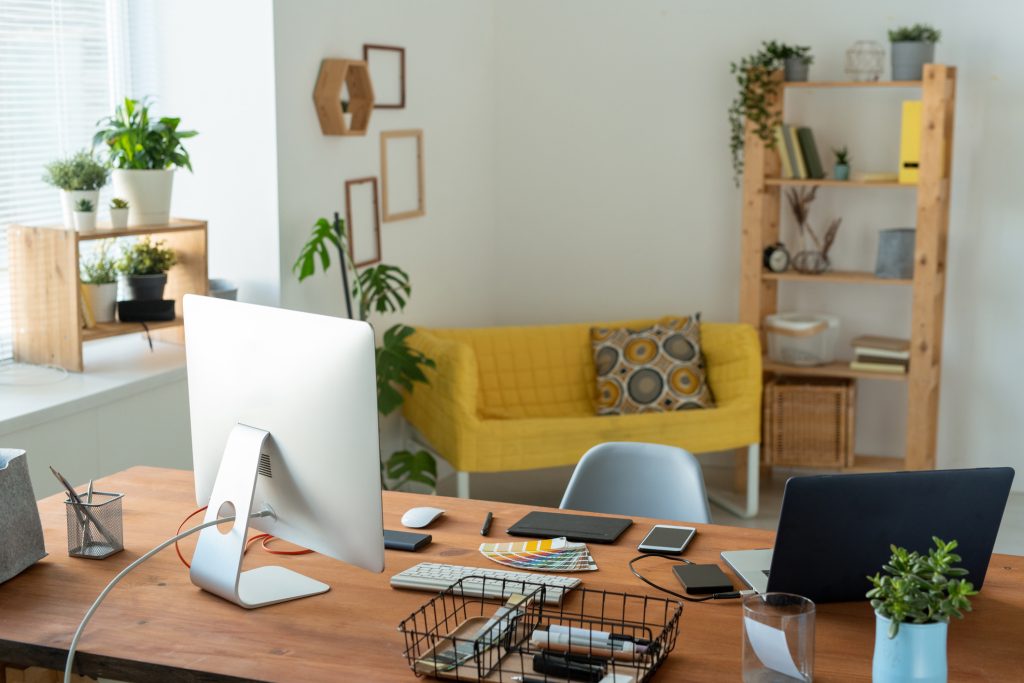 A home office where a desk with a computer monitor is placed perpendicular to the window. Tasteful furniture adds a splash of color in the background.
