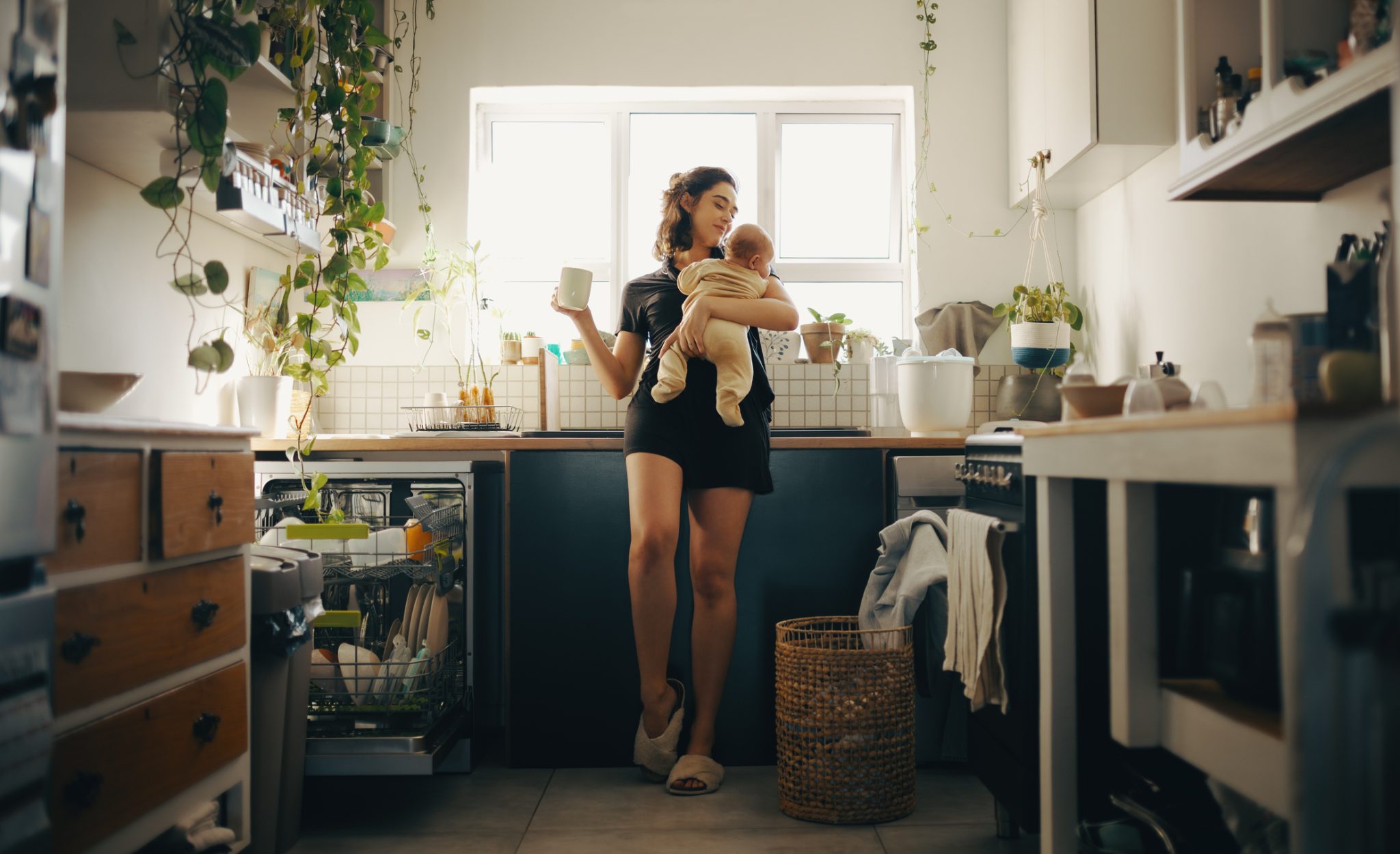 Woman with baby enjoying morning coffee in a bright, plant-filled custom home kitchen with natural light.