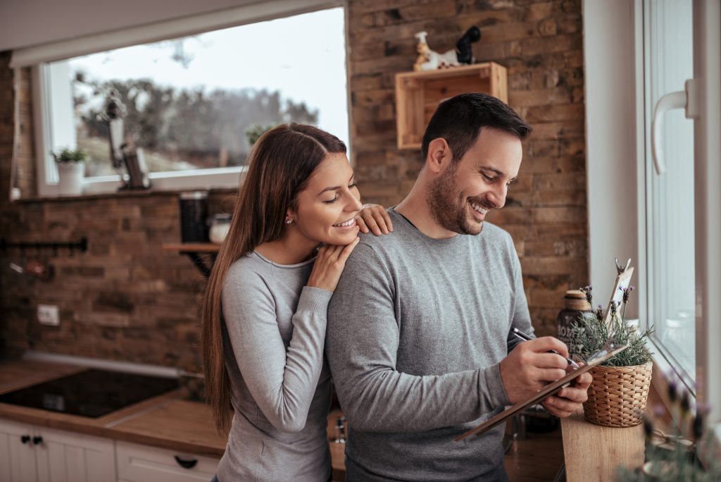 Millennial couple writing a home tour checklist together while planning to buy a house in their cozy apartment.