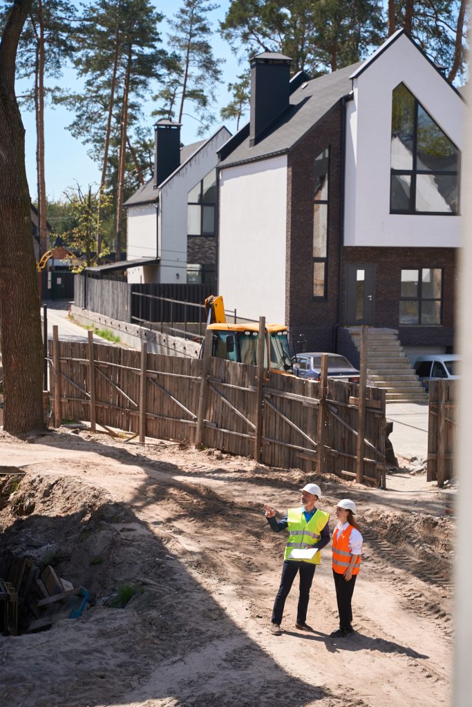 Home builder and homeowner standing at a fence, looking at an empty land lot next to a newly built custom home